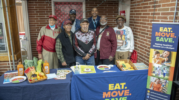 Group of participants and educators standing behind an Eat. Move. Save. table displaying nutrition education materials.