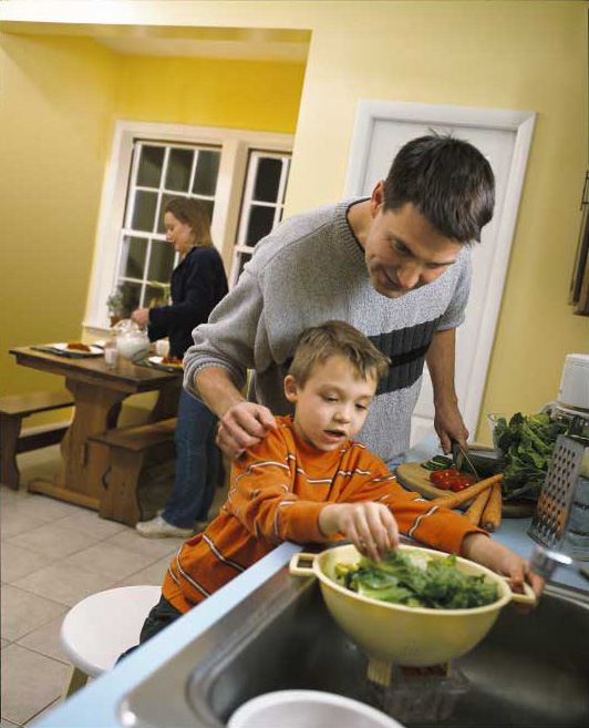 A father and son preparing a salad by the kitchen sink while the mother sets the table in the background