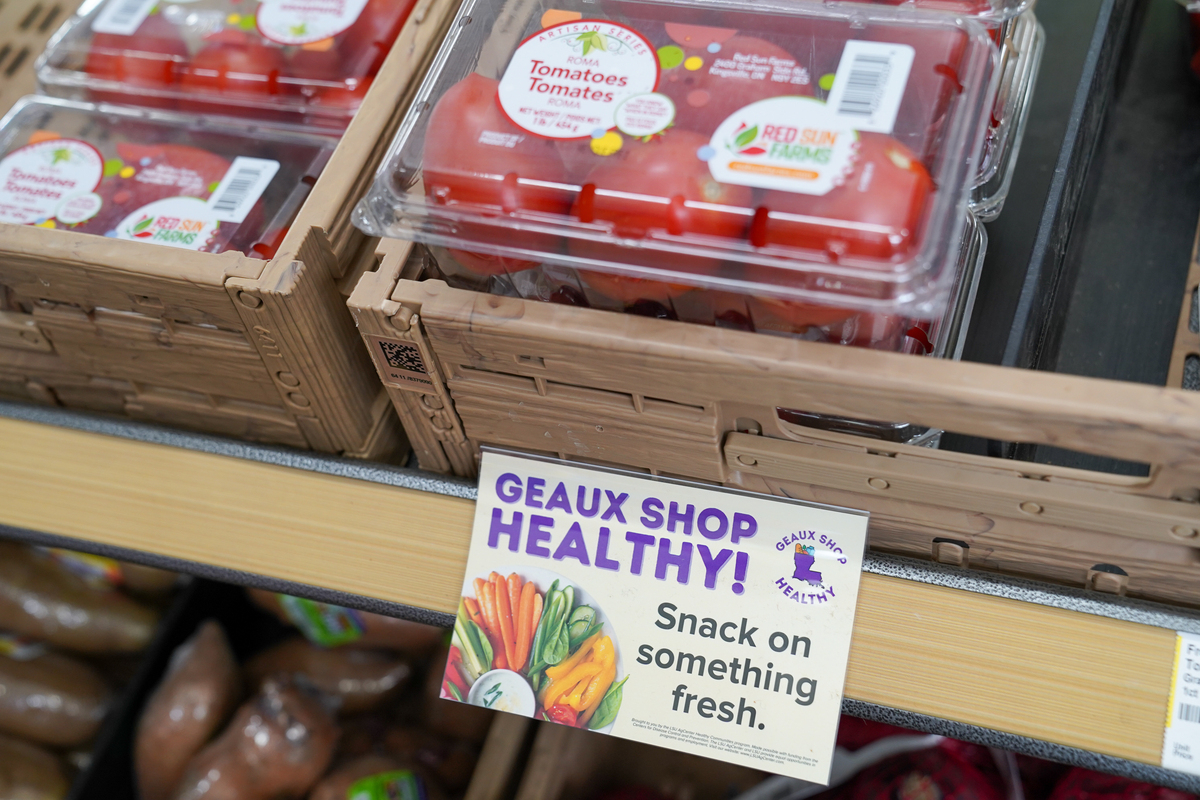 Tomatoes on a store shelf and a sign "Geaux Shop Healhty! Snack on something fresh"