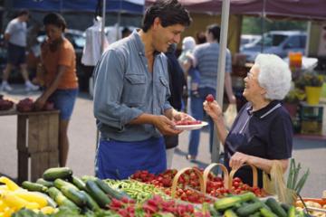 A grocer at a farmers' market offers an older woman a sample of strawberries
