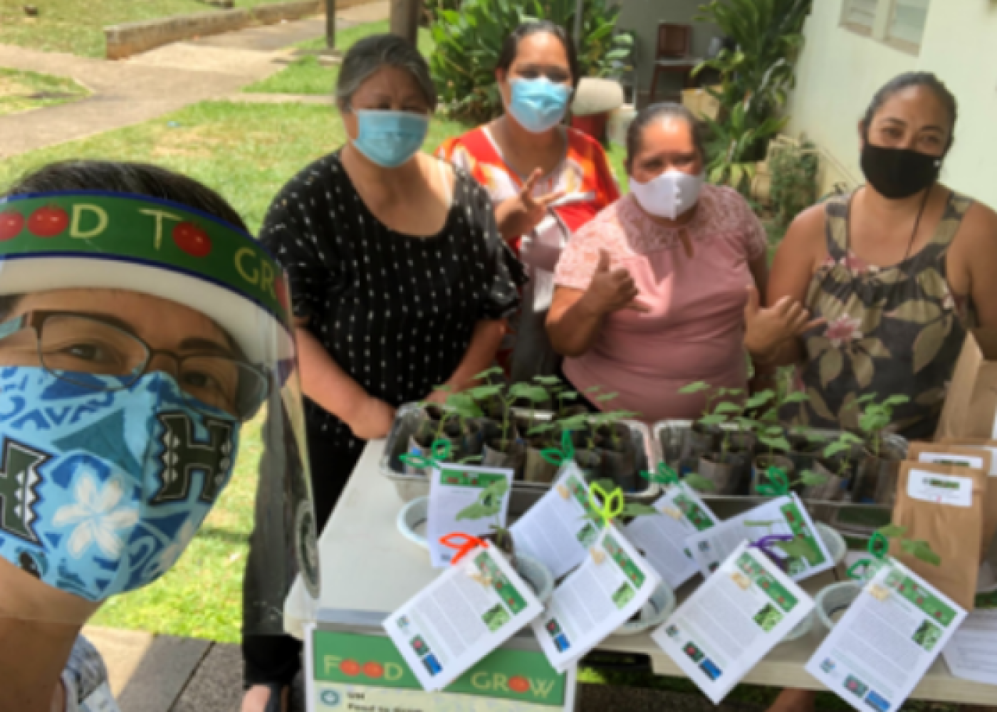 4 women standing behind their table of started vegetable plants with an educator in a "Food to Grow" COVID mask.