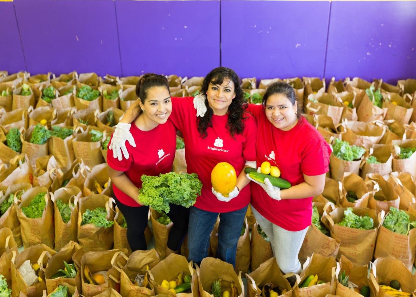 three women stand among a bunch of produce filled bags