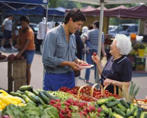 A grocer at a farmers' market offers an older woman a sample of strawberries