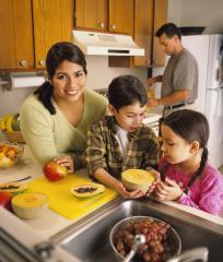 A mother and two children prepare fruit salad by the sink while father pours juice from a pitcher in the background